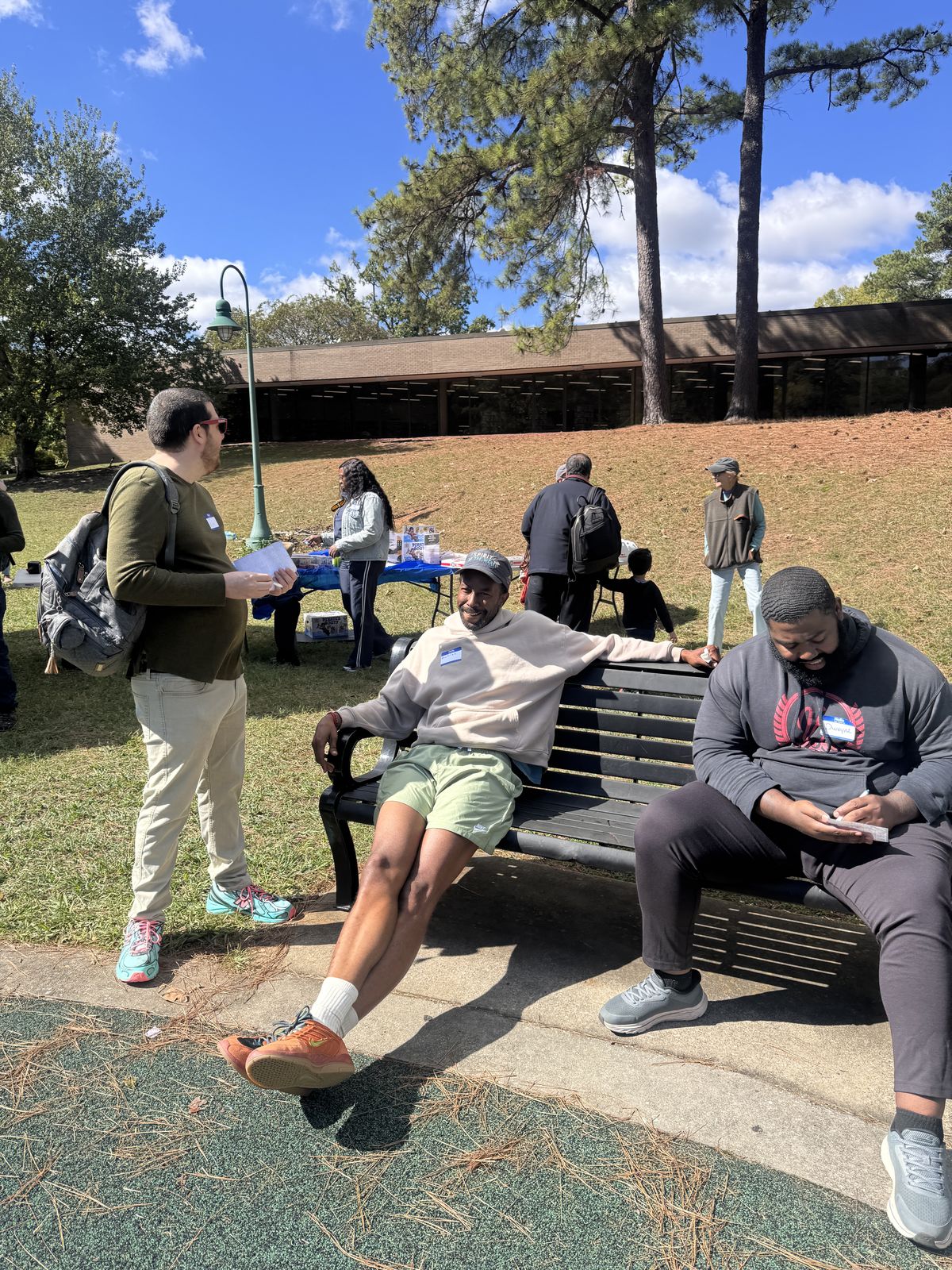 Three dads wearing name tags talking and laughing on and around a park bench on a sunny day.