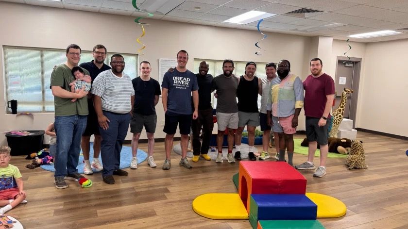 About a dozen dads standing together for a group photo in a playroom, one holding a baby, with kids playing nearby.