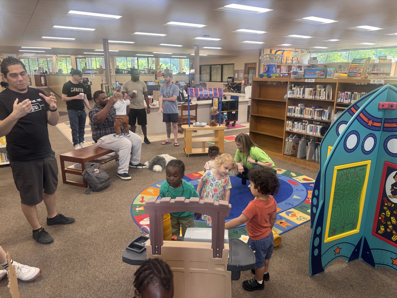 Dads holding coffee and chatting while toddlers play with toys in a library children's area.
