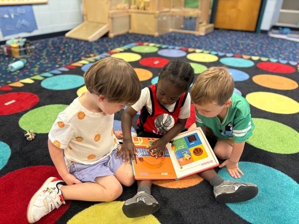Three toddlers sitting on a colorful rug, reading a board book about basketball together.