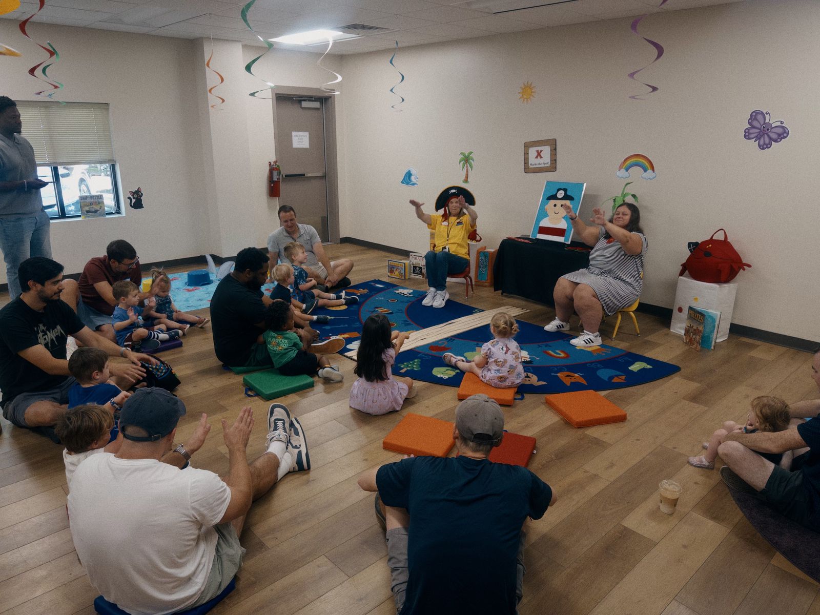 Dads sitting on the floor with their kids around a story-time rug while a woman in a pirate hat leads a song.
